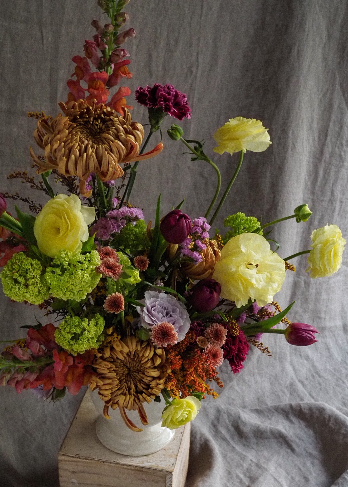 Colorful flower arrangement in a white vase on a wooden box with a gray fabric background